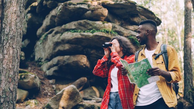 Happy brunette with curly hair is looking through binoculars standing in forest near rocks while her male friend is looking at map people are talking