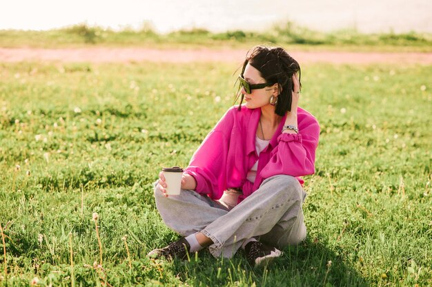 Happy brunette woman in casual clothes sitting on the lawn and resting in the park in sunny day