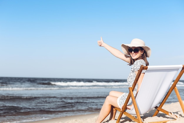 写真 親指を立てながら、海のビーチで木のデッキチェアに座っている幸せなブルネットの女性。
