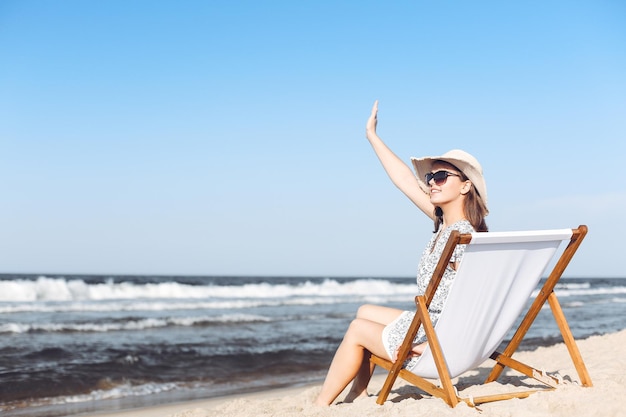 Счастливая брюнетка сидит на деревянном кресле на пляже океана, махав и приветствуя кого-то рукой.