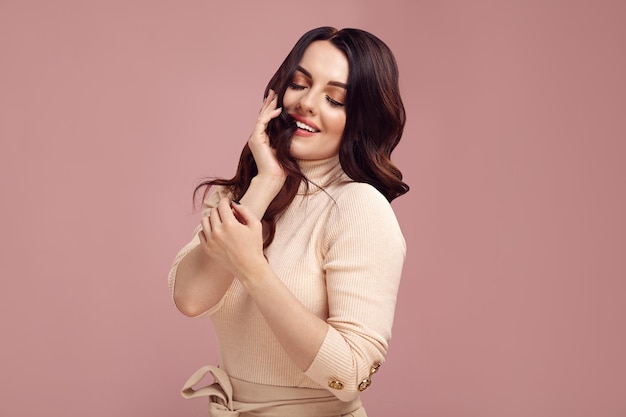 Happy brunette woman touches her hair and expresses satisfaction while standing isolated on a pink