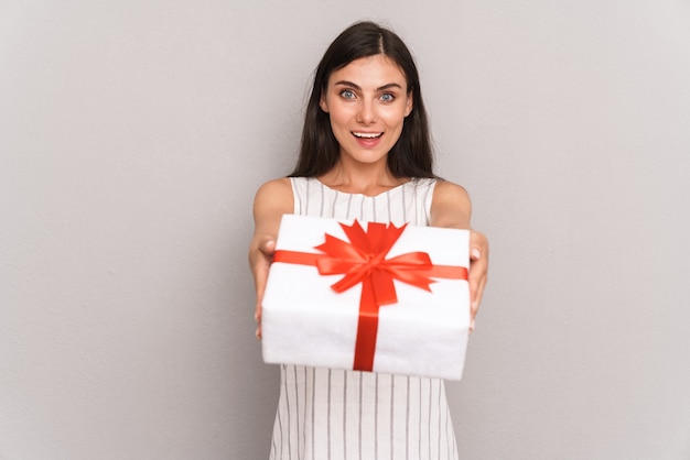 happy brunette woman wearing dress smiling and holding present box  isolated over gray wall