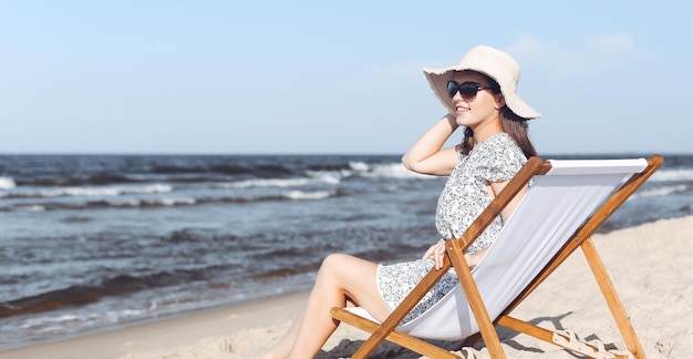 Happy brunette woman wearing sunglasses and hat relaxing on a wooden deck chair at the ocean beach.