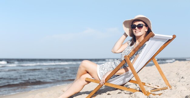 Happy brunette woman wearing sunglasses and hat relaxing on a wooden deck chair at the ocean beach.