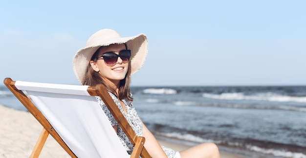 オーシャンビーチの木のデッキチェアでリラックスしたサングラスと帽子をかぶった幸せなブルネットの女性。