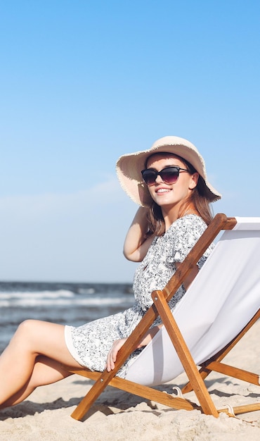 Счастливая брюнетка в солнцезащитных очках и шляпе расслабляется на деревянном кресле на пляже.