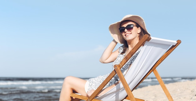 Happy brunette woman wearing sunglasses and hat relaxing on a wooden deck chair at the ocean beach