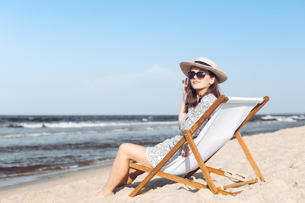 Happy brunette woman wearing sunglasses and hat relaxing on a wooden deck chair at the ocean beach.