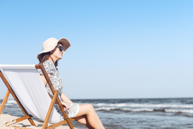 Happy brunette woman wearing sunglasses and hat relaxing on a wooden deck chair at the ocean beach.