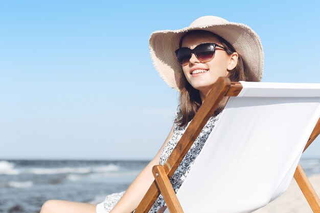 Happy brunette woman wearing sunglasses and hat relaxing on a wooden deck chair at the ocean beach.