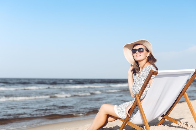 Happy brunette woman wearing sunglasses and hat relaxing on a wooden deck chair at the ocean beach.