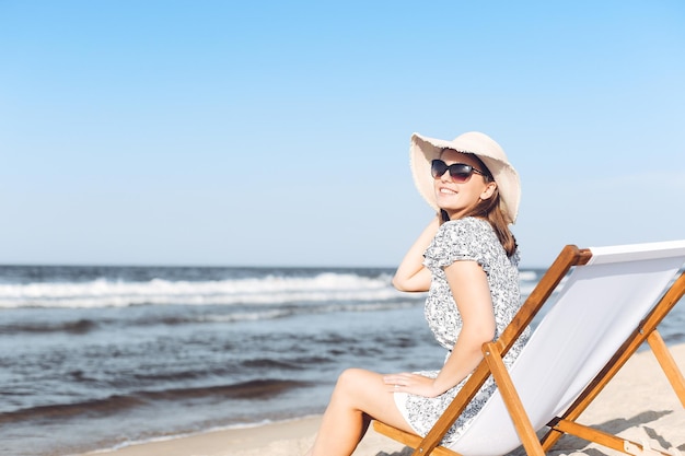 Happy brunette woman wearing sunglasses while relaxing on a wooden deck chair at the ocean beach