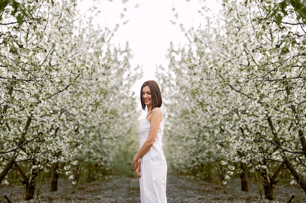 Happy brunette woman in white dress smiling while standing by blooming lane at spring park