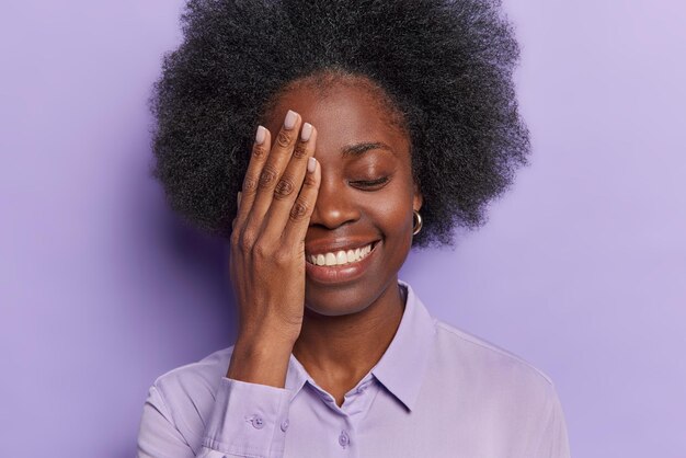 Photo happy brunette woman with curly bushy hair covers half of face keeps eyes closed has joyful expression stands carefree dressed in shirt isolated over purple background smiles sincerely feels upbeat
