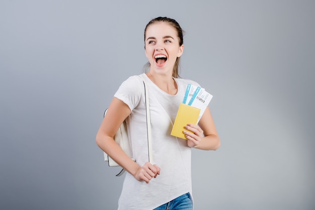 Happy brunette woman with plane tickets in passport and backpack isolated over grey