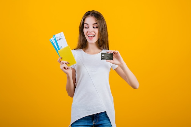 Happy brunette woman with plane tickets in passport and credit card isolated over yellow