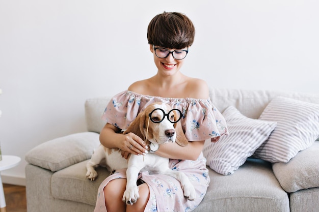 Happy brunette young woman and beagle dog in similar glasses having fun together and posing on gray background. Lovely lady with trendy haircut playing with puppy fooling around on sofa.