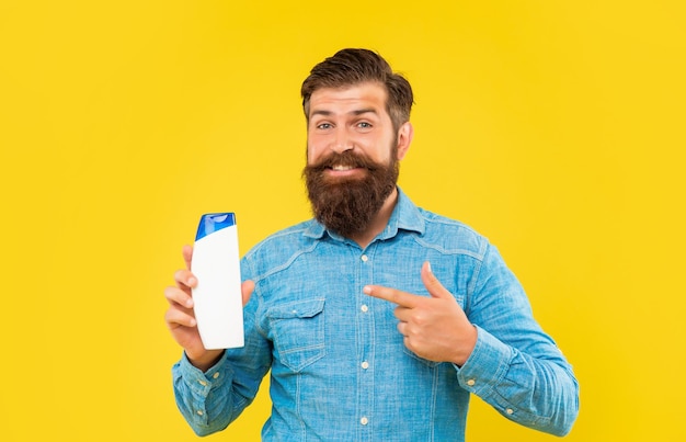 Happy brutal bearded man point finger on hair conditioner bottle on yellow background haircare