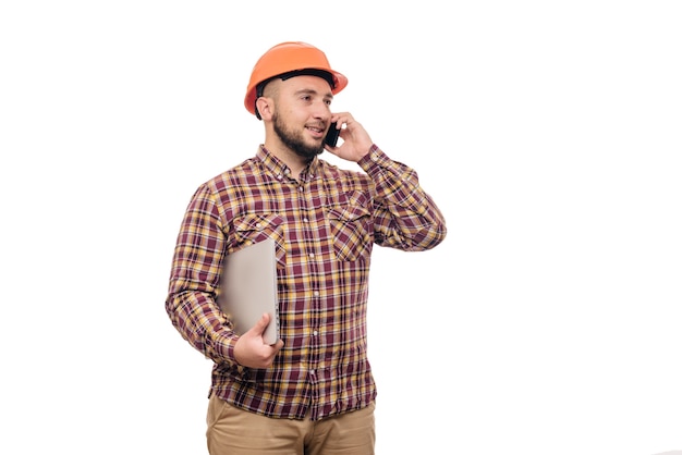 Operaio felice del costruttore nel casco arancione della costruzione protettiva che tiene un computer portatile e che parla sul telefono, isolato su fondo bianco. copia spazio per il testo. ora di lavorare.