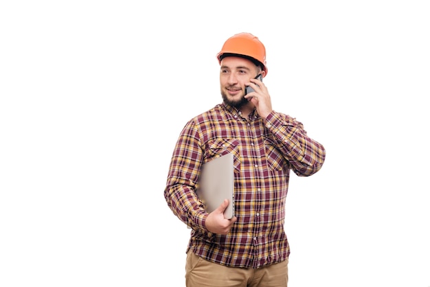 Foto operaio felice del costruttore nel casco arancione della costruzione protettiva che tiene un computer portatile e che parla sul telefono, isolato su fondo bianco. copia spazio per il testo. ora di lavorare.