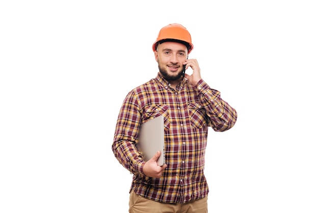 Foto operaio felice del costruttore nel casco arancione della costruzione protettiva che tiene un computer portatile e che parla sul telefono, isolato su fondo bianco. copia spazio per il testo. ora di lavorare.