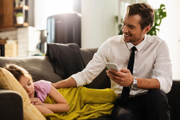 Happy business father sitting next to his sleepy daughter and text messaging on smart phone