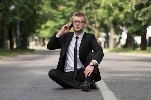 Счастливый деловой человек с помощью мобильного телефона на улице сидит на асфальте