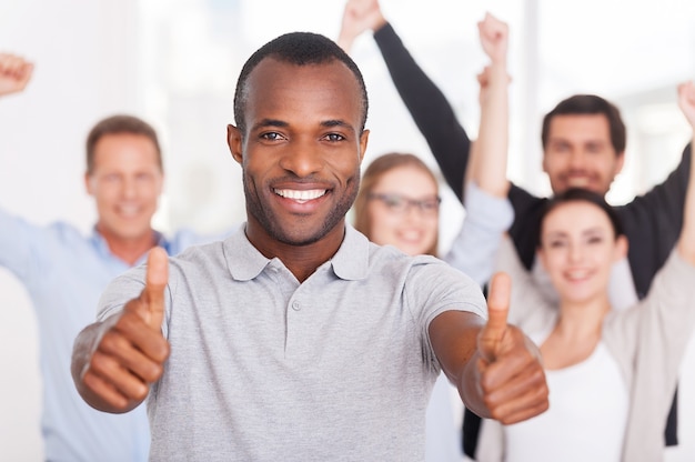 Happy business team. Happy young African man showing his thumbs up you and smiling while group of people in casual wear standing on background