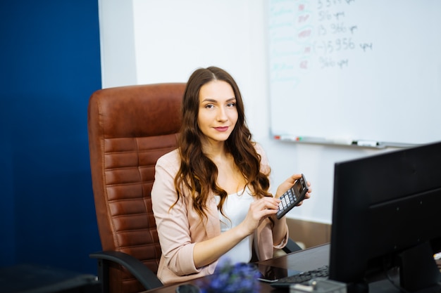 Счастливый бизнес женщина шатенка босс с длинными волнистыми волосами, улыбаясь, вдумчивый, сидя на офисном стуле в офисе и рядом с компьютером