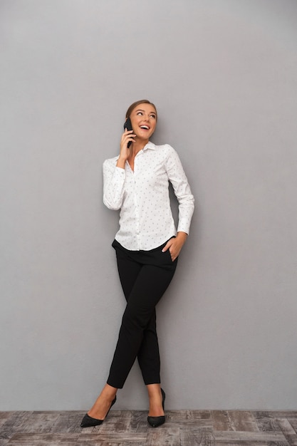 Photo happy business woman standing over grey wall background talking by mobile phone.