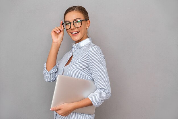 Happy business young woman posing isolated over grey wall background holding laptop computer.