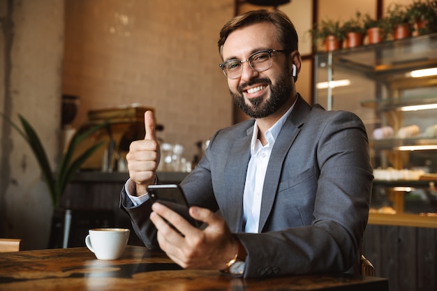 Happy businessman drinking coffee while sitting at the cafe table, holding mobile phone