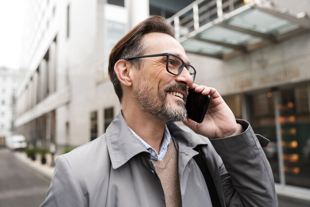 happy businessman in eyeglasses talking on cellphone and smiling while standing near office building at city street