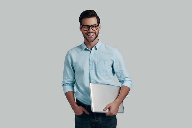 Happy businessman. Handsome young man carrying laptop and looking at camera while standing against grey background