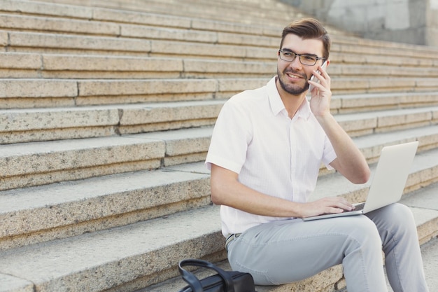 Happy businessman is having a pleasant talk on phone and working on laptop computer while sitting on stone stairs next to his briefcase, closeup, copy space. Technology, communication and business