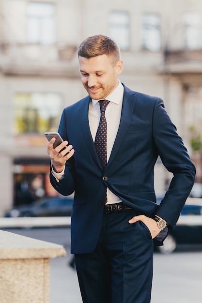 Happy businessman keeps hand in pocket wearing formal suit and wristwatch and using smart phone
