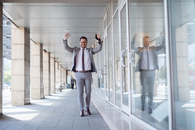 Happy businessman lifting arms while passing shop window. In one hand tablet and smart phone in other.