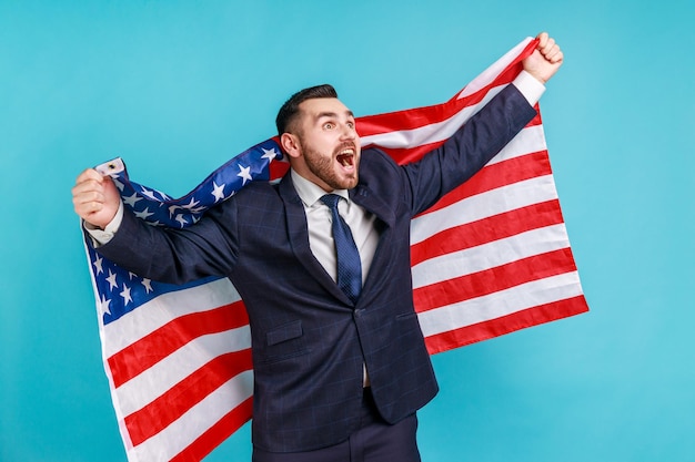 アメリカの独立記念日を祝う喜びのために叫んでアメリカの国旗を掲げるスーツを着た幸せなビジネスマン7月の政府のビジネスサポートの4日青い背景で隔離の屋内スタジオショット