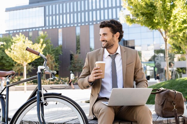 Happy businessman in suit using laptop and holding paper cup while sitting on bench near bike