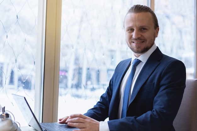 Happy businessman in the suit  working on the laptop sits and looking at the camera 