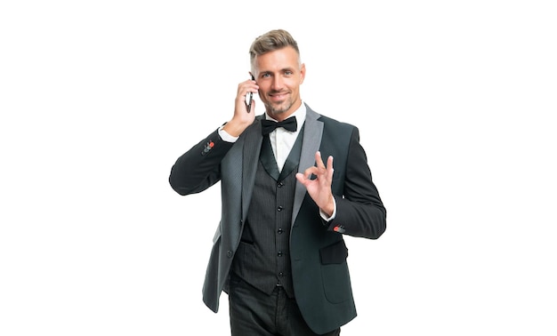 Happy businessman in tuxedo speak on phone isolated on white background show ok