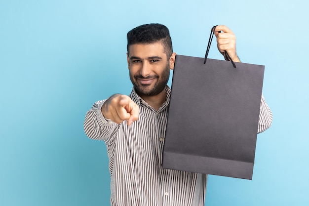 Happy businessman with beard pointing finger at camera holding black paper bag with purchases