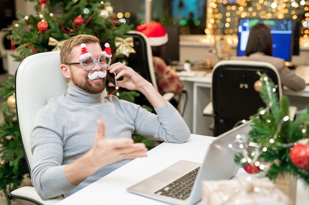 Photo happy businessman in xmas eyeglasses congratulating business partners on the phone in front of laptop in office