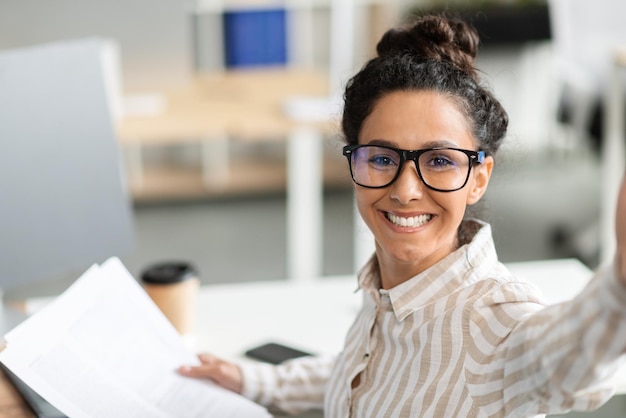 Happy businesswoman having video call or taking selfie while working with documents in office and smiling at camera