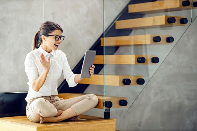 A happy businesswoman in lotus position sitting on the stairs and having video call on tablet