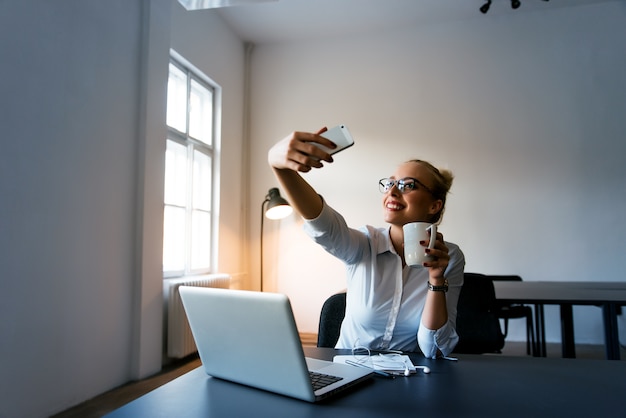 Foto donna di affari felice che fa selfie in ufficio con la tazza di caffè