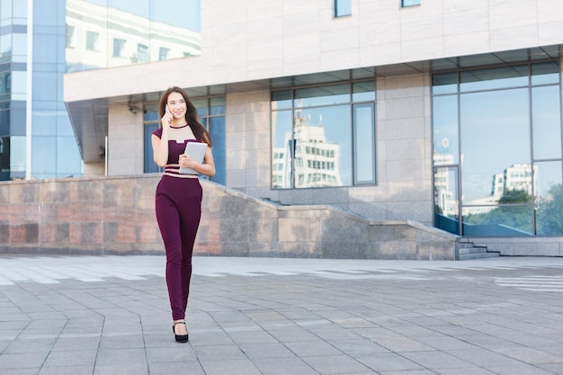 Happy businesswoman talking on mobile and working with tablet. Woman holding digital device and consulting on smartphone, standing near modern office center, copy space