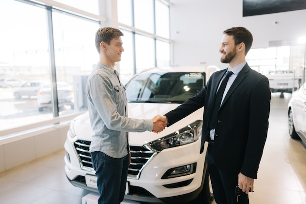 Happy buyer of car shaking hands with seller in auto dealership in front of car