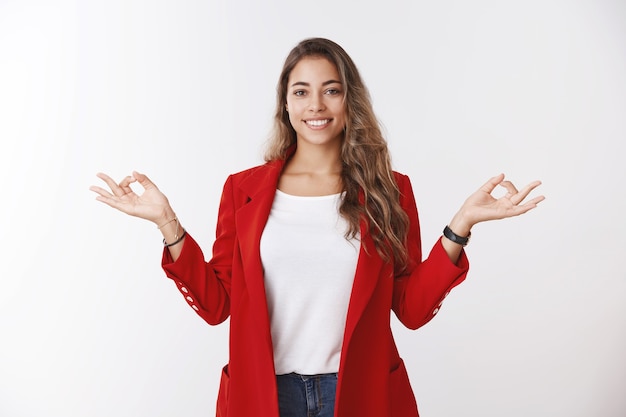 Happy calm stress-free modern 25s businesswoman control feelings hands sideways showing mudra zen gesture smiling peaceful relieved, meditating doing yoga wearing office red jacket