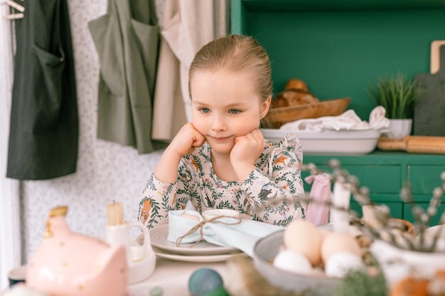 Happy candid little kid four year old girl bored with hands fists in face ready and waiting for lunch or dinner of springtime Easter holiday at home kitchen decorated festive table with Easter eggs